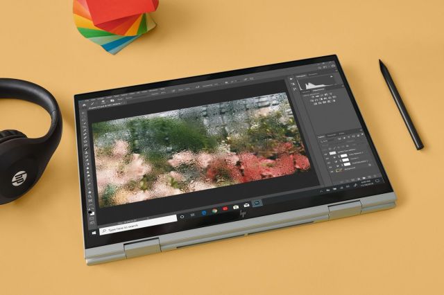 HP’s Envy x360 15 (Intel) in tent mode on a yellow background seen from above. To its right is a pair of headphones and to its left is a stylus. Above it is a stack of multicolored post-it notes. The screen displays a photo-editing software. 