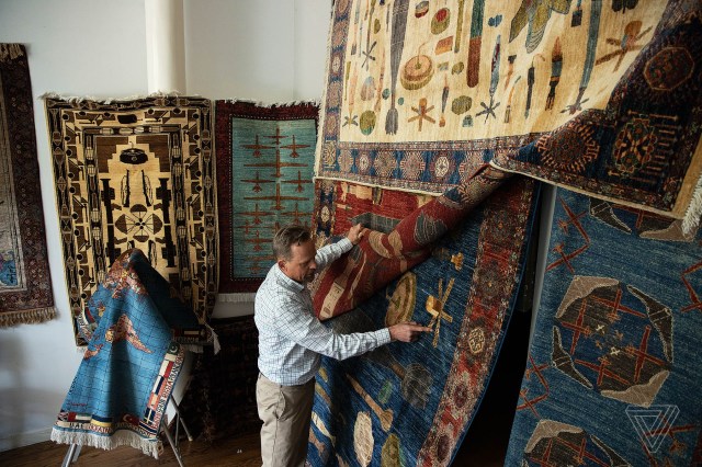 Sudeith with large war rugs in his apartment. He pulls aside a red rug to point to out details on a blue rug beneath it.
