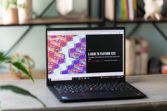 The Lenovo ThinkPad X1 Carbon Gen 9 on a table in front of a bookshelf, open, angled slightly to the right. The screen displays a Verge article.