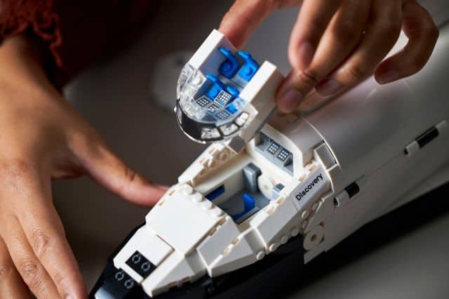A hand lifting the flight deck from the space shuttle, revealing another deck underneath. Both decks have tiny blue chairs and tiny control panels.