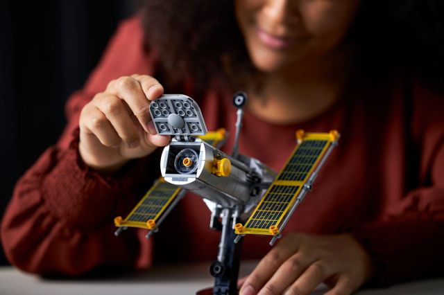 A person lifting a little door at the end of the Hubble telescope, showing a tiny clear piece that represents the telescope’s lens. 