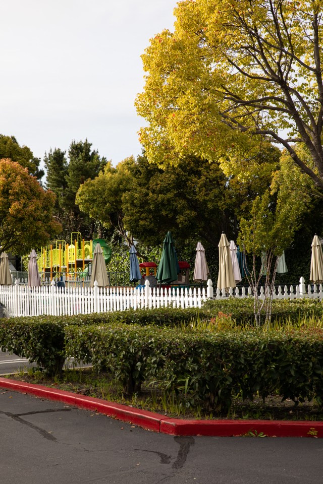 At the edge of a parking lot lush trees and shrubs sandwich a white fenced in area of tables with closed umbrellas. A brightly painted playground is behind it.