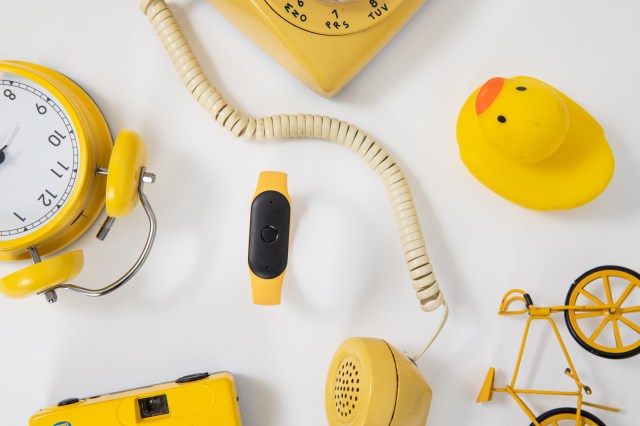 The Bee AI wearable in the yellow wristband surrounded by several yellow objects, such as a telephone, alarm clock, camera, rubber duck, and a toy bicycle.