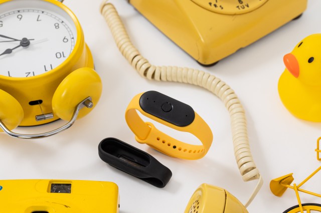 Side angle of Bee wearable surrounded by yellow objects, including a yellow alarm clock, yellow rotary phone, rubber duck, and bicycle. To its lower left, you can see the black pin attachment.