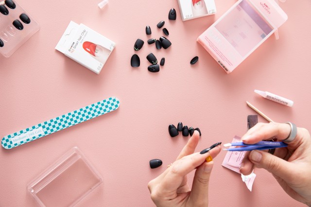 Top-down view of person applying Jakcom N3 Smart Nail chip to their nail. The supplies are scattered about, including a nail file, lots of matte black nails, empty boxes, cuticle stick, alcohol pad, and nail glue.