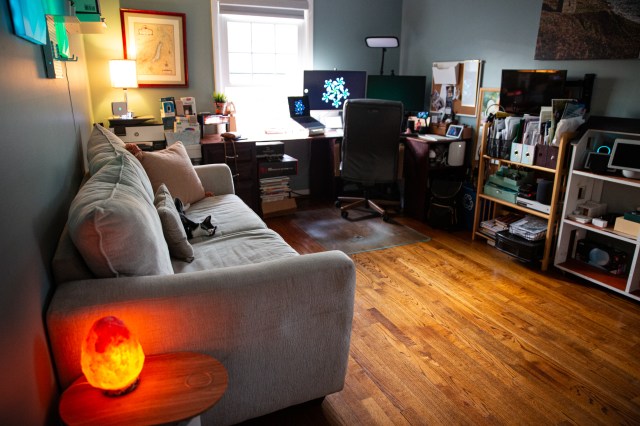 A home office with a couch on the left, a window at the far end, and a corner desk filled with tech.