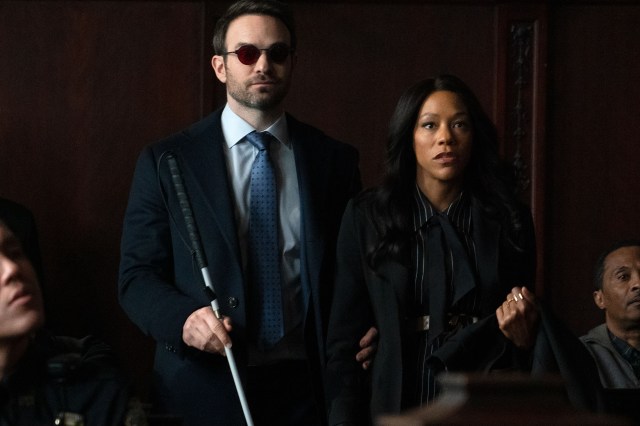 A blind man in a business suit standing next to a woman in a suit. The pair are standing in a courtroom.