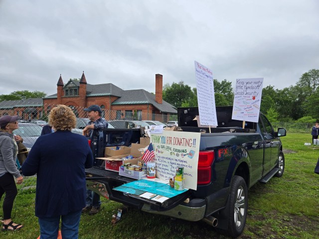 Three people standing around a pickup truck with a food bank donation sign. A protest sign behind it reads “keep your nasty little hands off social security.”