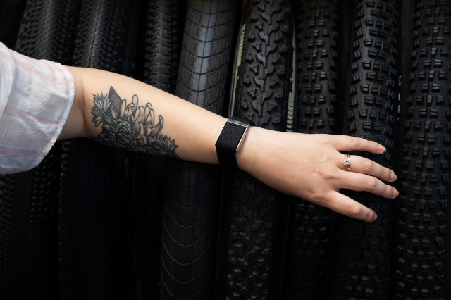Person with a floral tattoo on the forearm browsing through a bike tire rack while wearing a short sleeved pink-and-white blouse and a Whoop MG on the wrist.