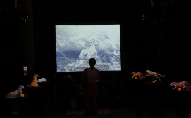 A dark room with a bright screen showing a digital jaguar. Several small jaguar statues surround a viewer in the center.