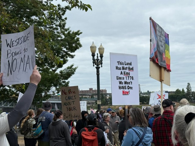 A crowd of protesters holding signs. One reads “Westside bows to no man,” another “This land has not had a king since 1776. We won’t have one now,” and another “No one cares about your birthday.”