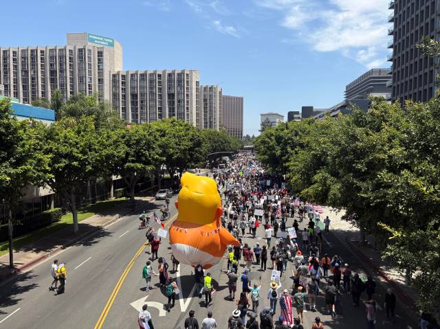 A parade balloon depicting Donald Trump as a baby wearing a diaper floats above a crowd of people marching down a street.