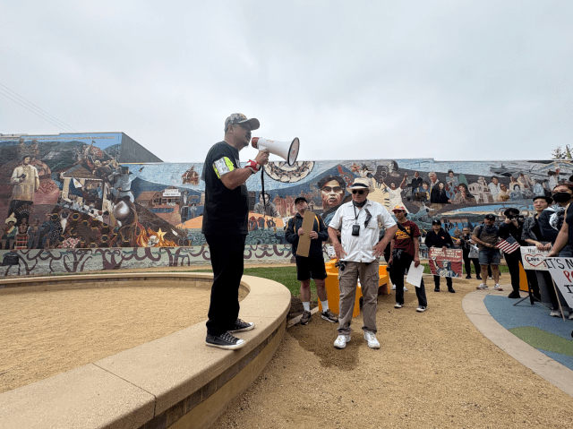 A man wearing a camouflage-patterned hat and a black t-shirt that says “No Kings in America” speaks to people at a park in front of a mural.