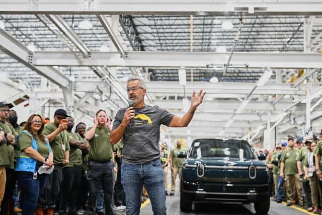 A man in a grey t-shirt and jeans holds a microphone and stands in front of a blue Rivian r2 inside of a huge, bright factory space and speaks to a crowd of people who are mostly wearing green t-shirts.