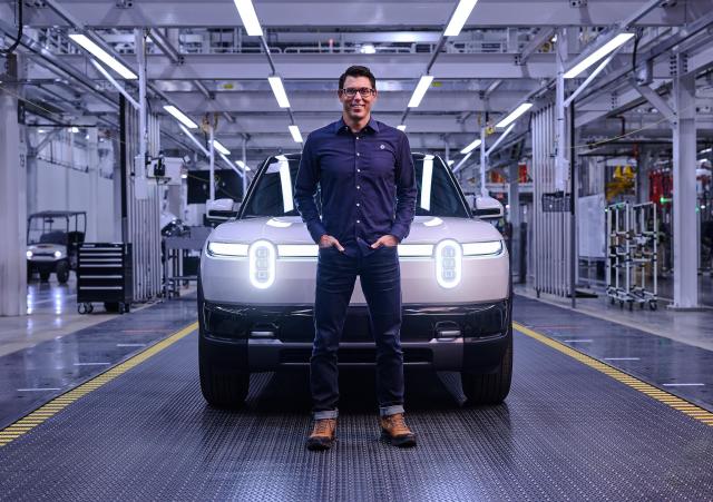 A smiling man wearing jeans, a blue button down shirt, boots and glasses stands in front of a white Rivian R2 with its headlights on. The image is taken in a factory and has a blue cast.