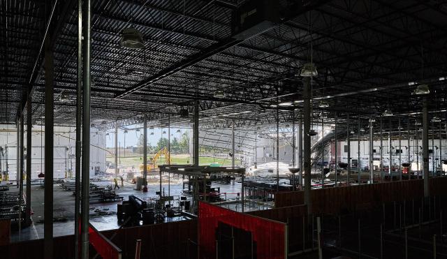 A wide view of a factory interior is mostly dark with a large open wall in the back where you can see to the bright daylight outside. A construction vehicle is in the center and debris can be seen hanging down. 