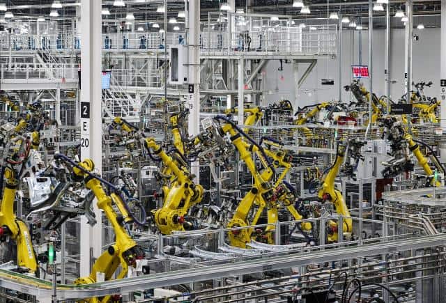 A wide and high view of the factory floor where yellow machine arms are seen lifting and working on unspecified car parts.