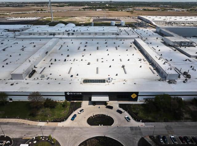 An aerial view of Rivian’s huge factory with rural land seen behind it and cars parked in its circular driveway in front. 
