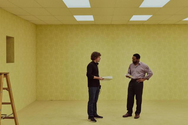 A image of two men on a film set made to look like the interior of a bland, beige office building.