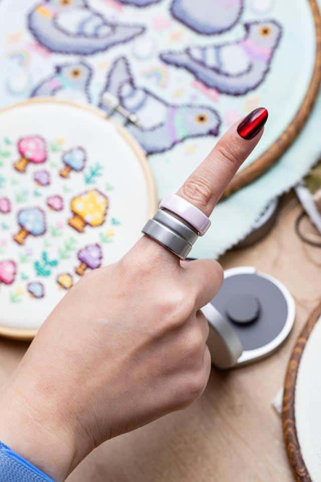 Someone wearing two Oura Ring 4s with an Oura Ring 5 in beween with cross stitch projects in the background.