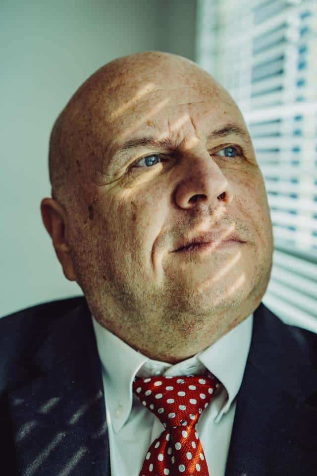 Buddy Rizer, an older man wearing a suit, poses for a portrait near the window in his office.