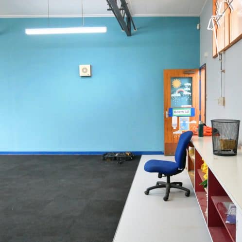 Bright blue wall inside a modern classroom with a door labelled "Room 10", a blue office chair, and a white shelf with colourful storage bins, creating an inviting learning environment.