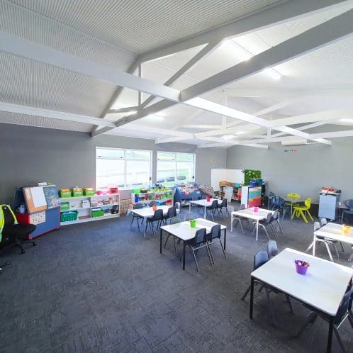 Bright indoor children's learning and activity room with tables, chairs, storage shelves, and educational materials, ideal for early childhood education and kid-friendly spaces.