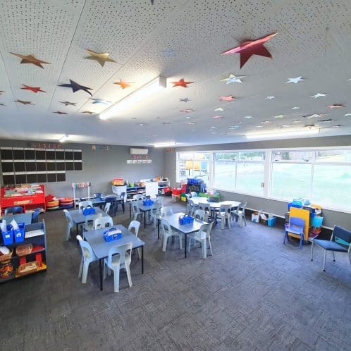 Bright and colourful early childhood classroom with tables, chairs, and educational materials, featuring star-themed ceiling decorations and large windows providing natural light.