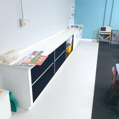 Clean classroom kitchenette with black storage cupboards, white countertop, and instructional safety posters, suitable for early childhood education or preschool settings.