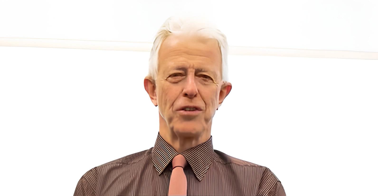 Old man with white hair in a brown shirt and ribbon tie, speaking or presenting, with a plain white background, professional business or corporate environment.