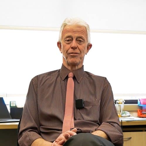 Elderly man in a brown dress shirt and pink tie sitting in an office setting, engaging in a discussion or presentation.