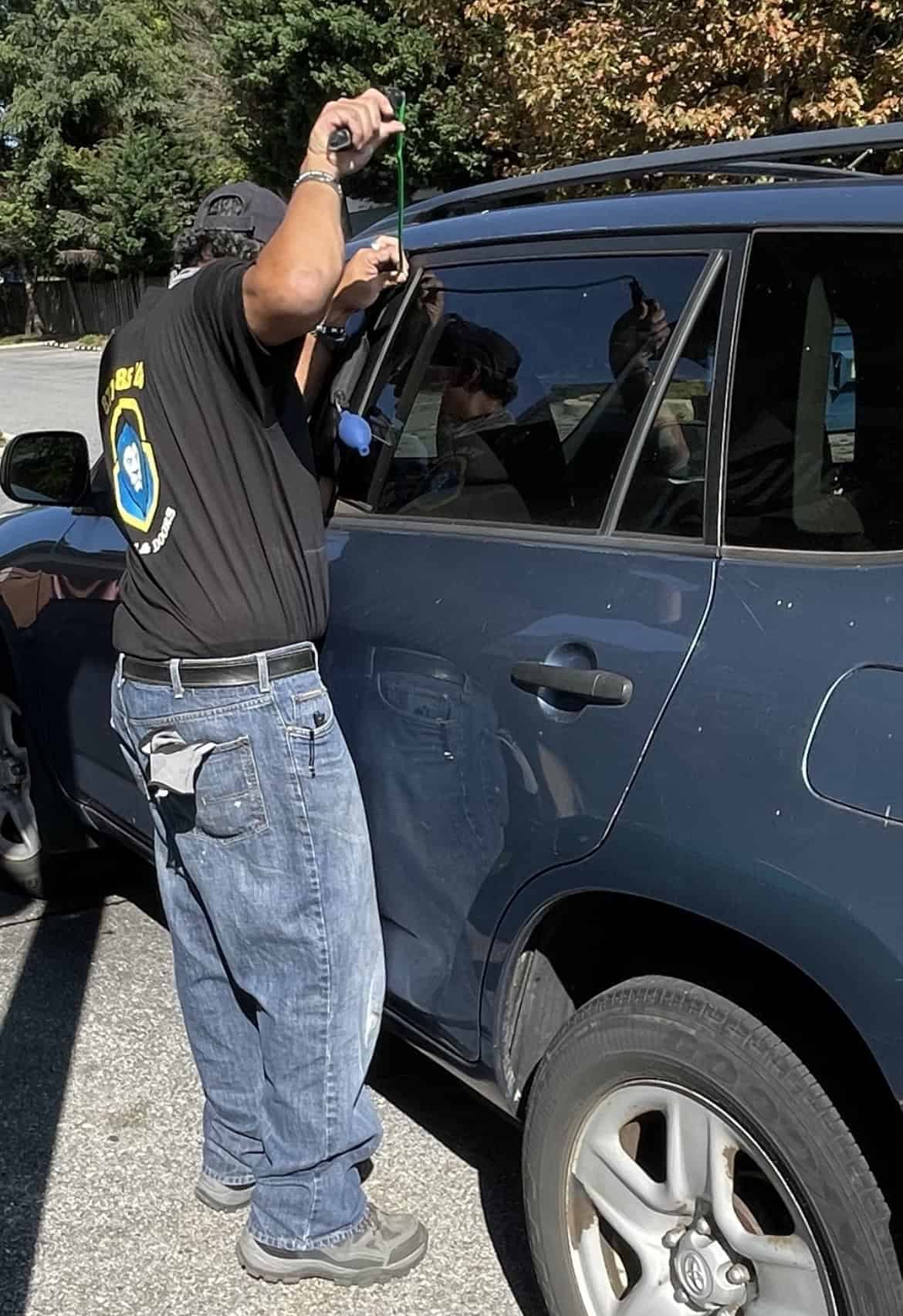 A Man Wearing A Black Locksmith Uniform Sits Standing In A Black Car Who Is An Auto Locksmith Expert