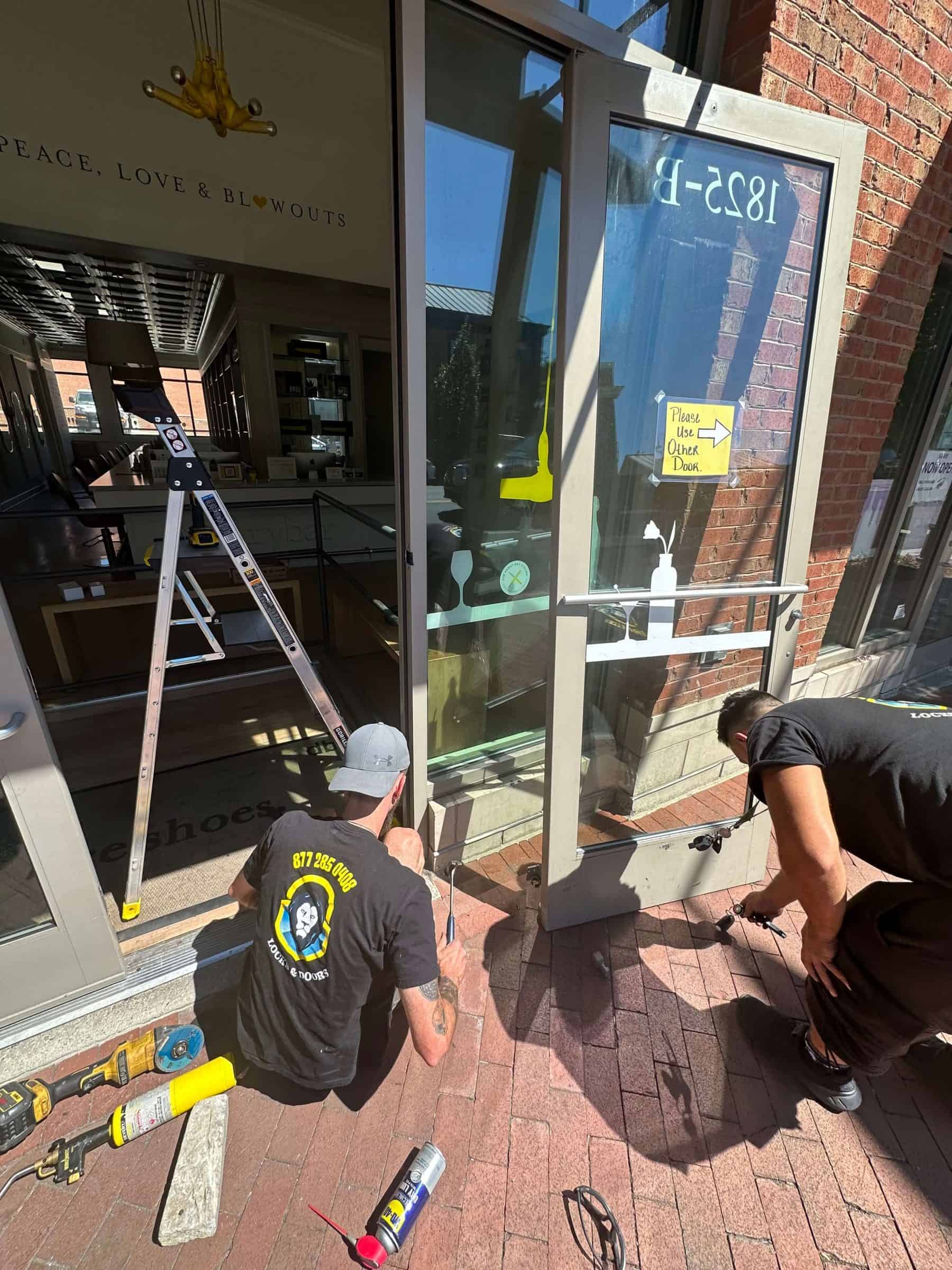Three Locksmiths Working On A Glass Door, With Tools And Equipment Spread Around Them.