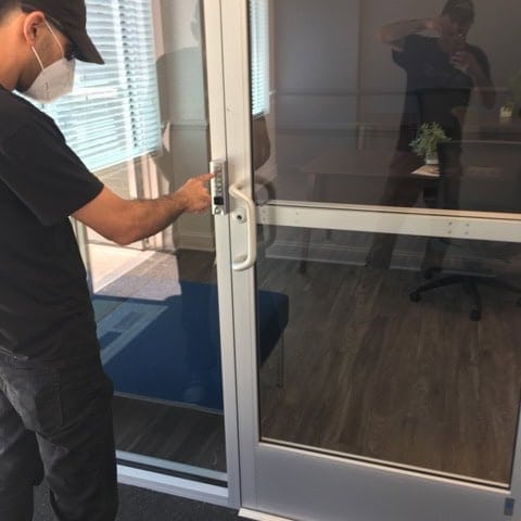A Locksmith Bethesda Md, Dressed In A Black Uniform And Wearing A White Mask, Inspecting The Door Knob.