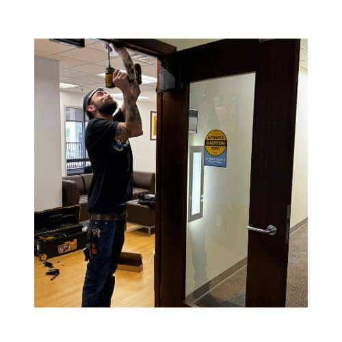 Locksmith From King Locksmith Installing An Automatic Door System On A Glass-Paneled Wooden Door In A Commercial Building In Burtonsville Md, Using A Power Drill, With Tools And Equipment Visible In The Background.