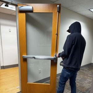 Fort Washington Md Locksmith Services Emergency Locksmith Fort Washington - Locksmith In A Hoodie Adjusting A Wooden Door With A Push Bar In A Commercial Interior Emergency Locksmith Fort Washington - Locksmith In A Hoodie Adjusting A Wooden Door With A Push Bar In A Commercial Interior