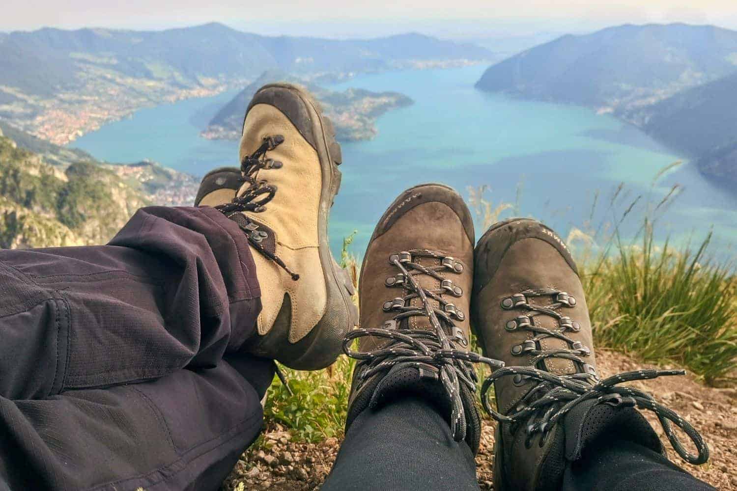 Two pairs of hiking boots rest at the edge of a mountain, overlooking a scenic lake and green hills in the distance under a clear sky.