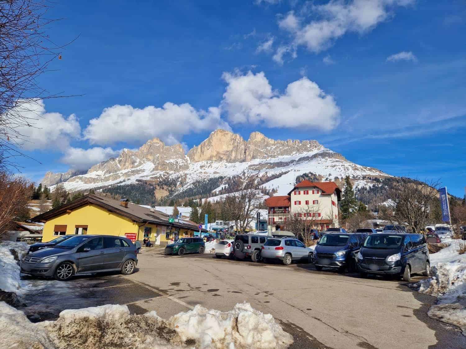 A snowy mountain landscape with cars parked in a lot, a yellow building, and several houses. The sky is blue with scattered clouds, and snow covers the ground and mountain slopes in the background.