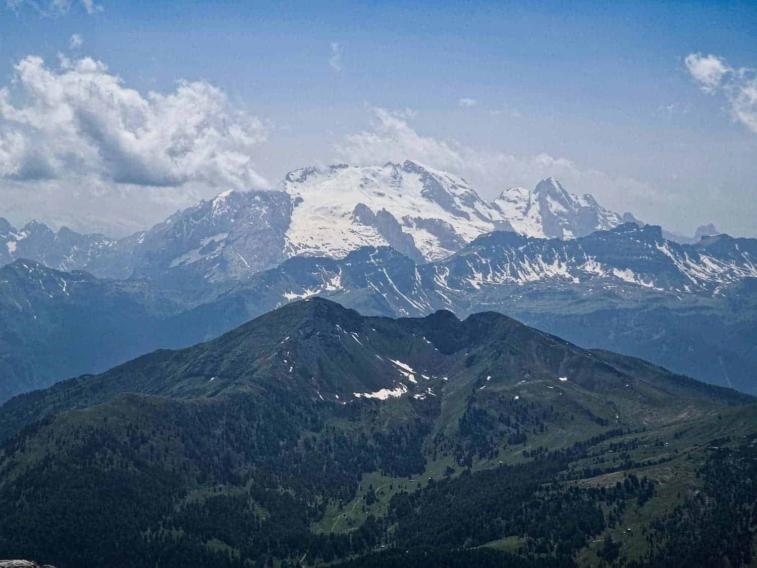 View of snow-capped mountains under a blue, cloudy sky. The foreground features grassy and rocky slopes leading to a distant, expansive range with prominent white peaks.