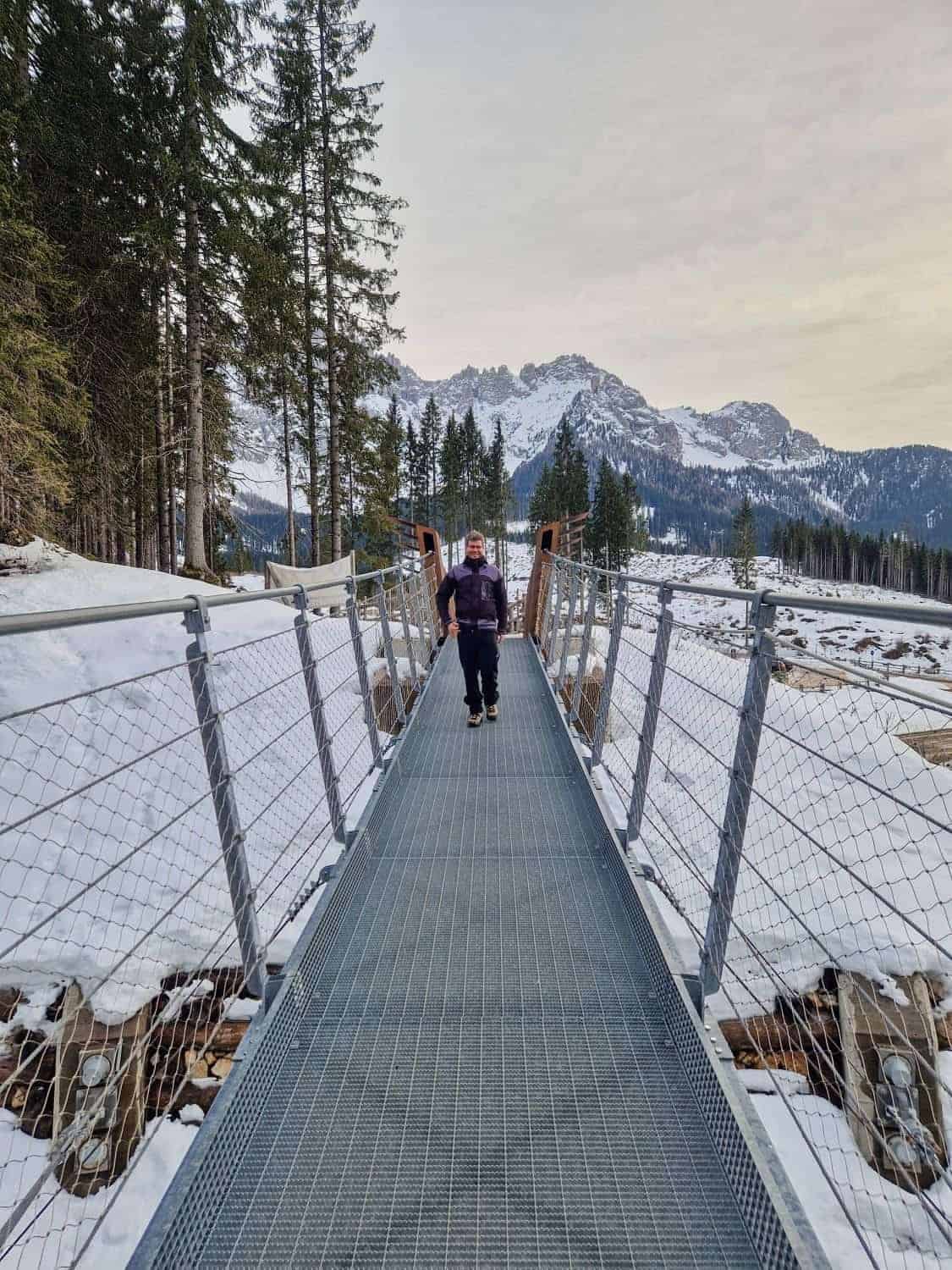 A person walks across a metal suspension bridge surrounded by snow-covered terrain and pine trees near Lake Carezza. Mountains with a light dusting of snow are visible in the background under an overcast sky.