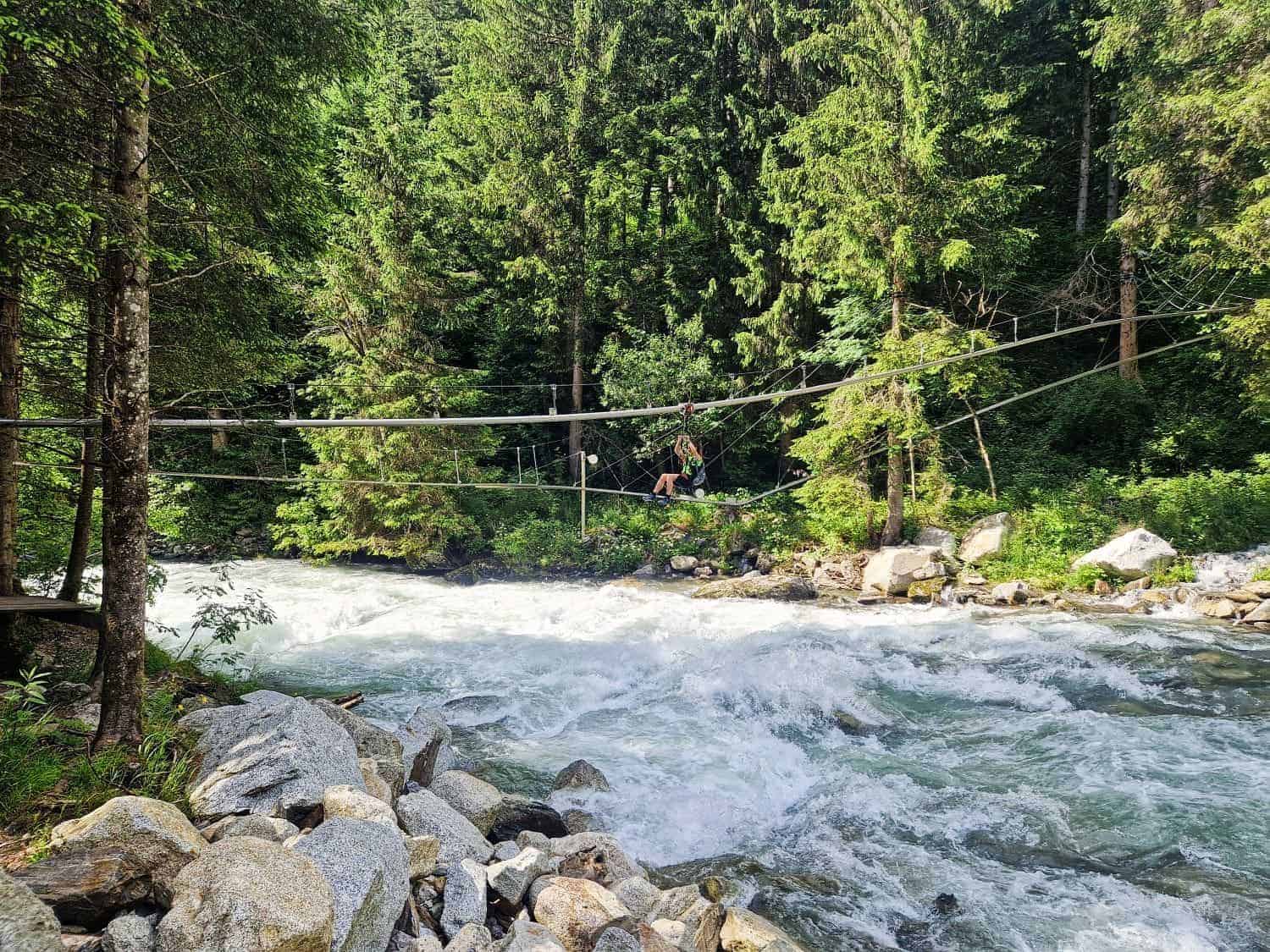A person crosses a rushing river using a zip line, surrounded by dense forest. Large rocks line the riverbank, and tall trees fill the background, creating a lush, natural setting.