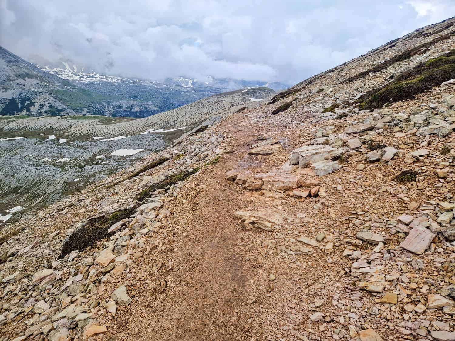 A mountain trail made of loose rocks and dirt winds through a rocky landscape with patches of snow. The overcast sky and scattered clouds suggest cool weather. Jagged peaks and valleys can be seen in the background, offering glimpses reminiscent of the stunning Cadini di Misurina viewpoint hike.