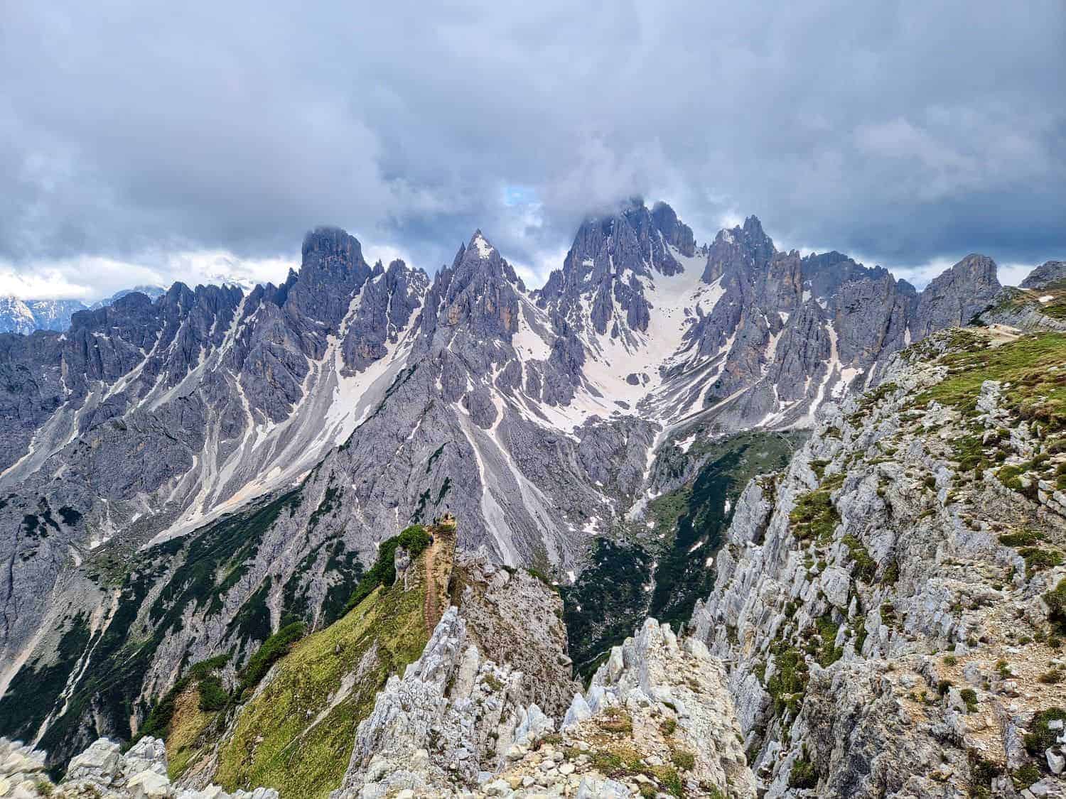 A rugged mountain range with jagged, snow-capped peaks under a cloudy sky. The Cadini di Misurina viewpoint hike reveals sharp cliffs and ridges amid patches of green vegetation. In the foreground, a rocky trail winds through the terrain, offering breathtaking vistas at every turn.