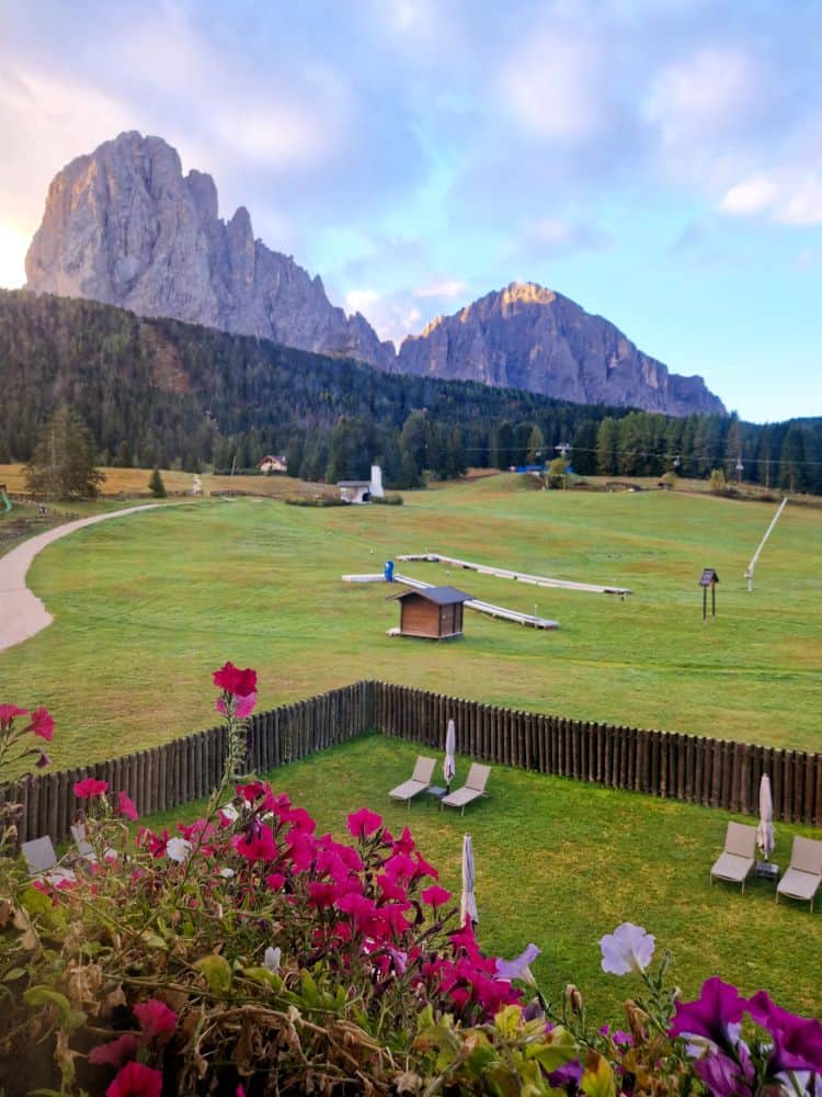 A scenic view of a grassy field with lounge chairs and umbrellas, bordered by a wooden fence and colorful flowers in the foreground, and dramatic mountains and forest in the background under a partly cloudy sky.