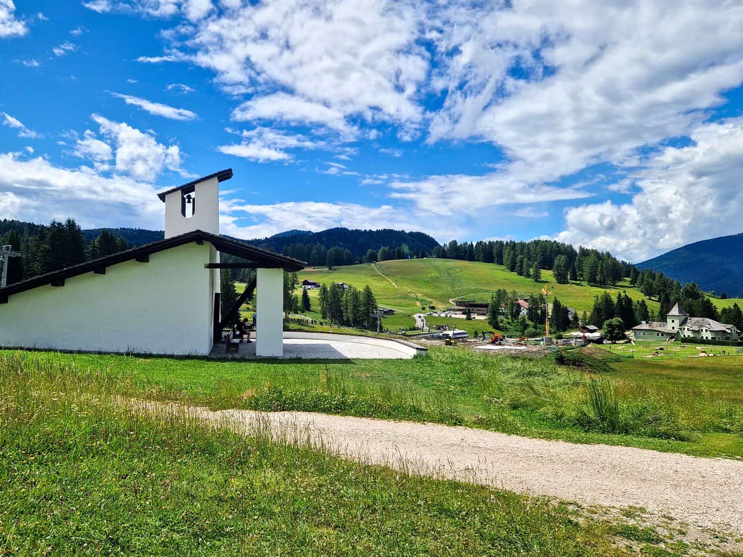 A small white chapel stands on a grassy hill under a partly cloudy sky, with a gravel path in the foreground and a village and forested hills in the background.