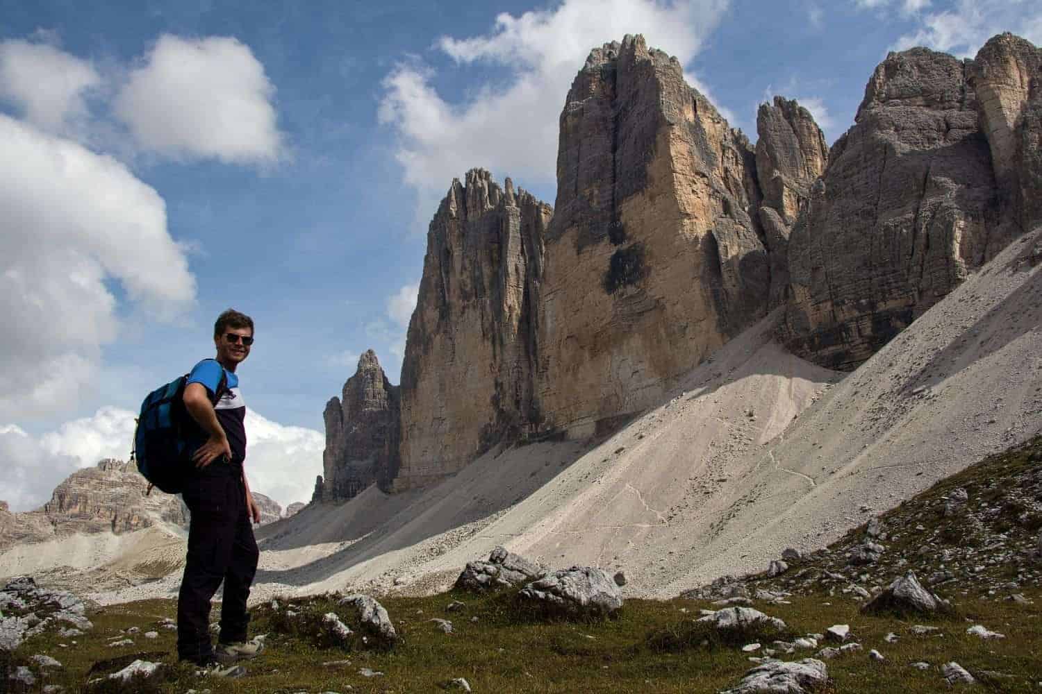 A hiker with a blue backpack stands on a grassy area, looking towards towering rock formations in the background under a partly cloudy sky. The scene suggests a rugged, mountainous landscape, possibly during a day hike or trek.