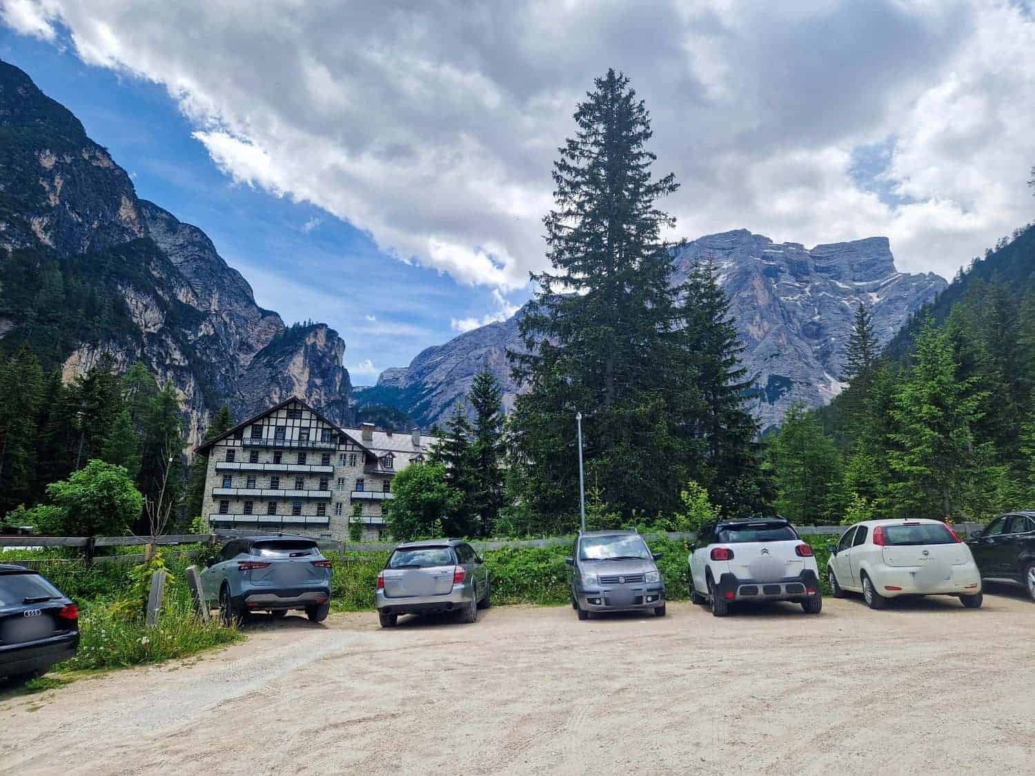 A parking lot with several cars is in the foreground. Behind, there are tall evergreen trees and a rustic building. In the background, majestic mountains rise under a cloudy sky, creating a picturesque natural setting.
