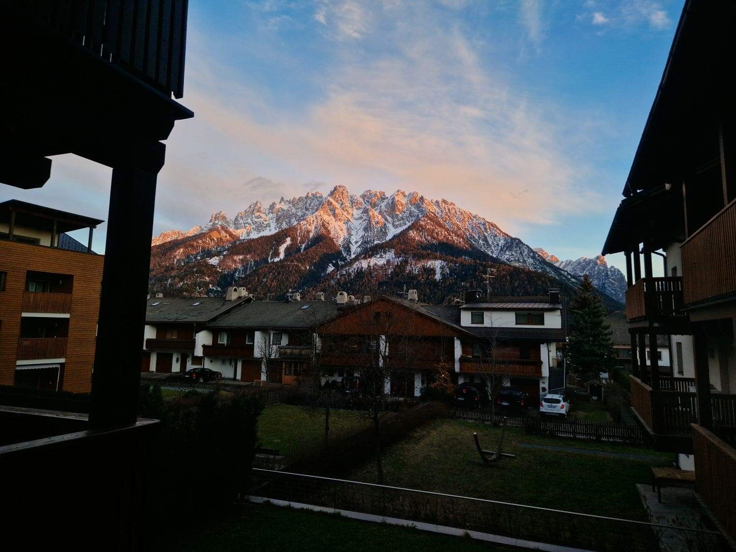 A scenic view of snowy mountains during sunset, with orange and pink hues reflecting on the peaks. In the foreground, several houses and buildings are silhouetted against the vibrant sky.