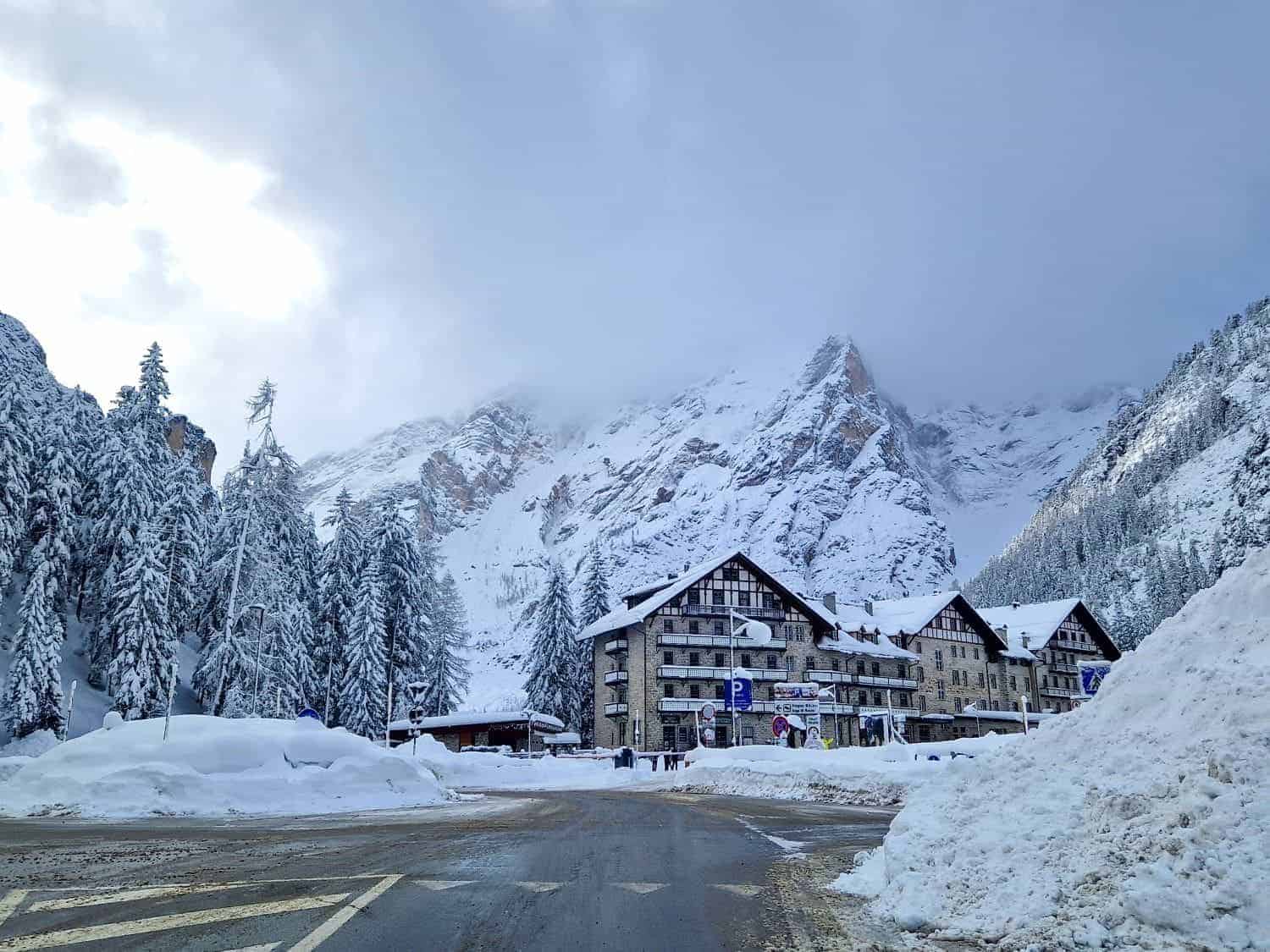 A snowy mountain scene with snow-covered buildings and trees. The sky is overcast, and mist partially obscures the mountain peaks in the background. The road in the foreground is clear with visible snow piled on the sides.