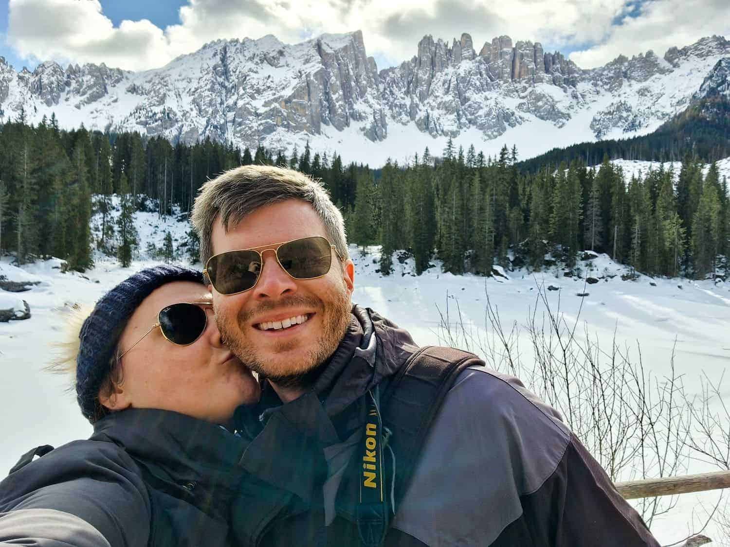 A couple dressed in winter jackets pose for a selfie in a snowy landscape. The woman kisses the man's cheek. Behind them are snow-covered trees and a mountain range under a partly cloudy sky. The man has a camera strap around his neck.